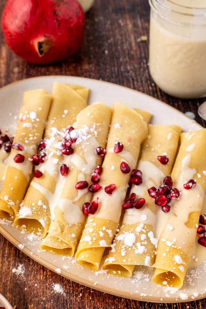 Close-up of rolled eggnog crepes drizzled with eggnog sauce, sprinkled with powdered sugar, and topped with bright pomegranate arils.