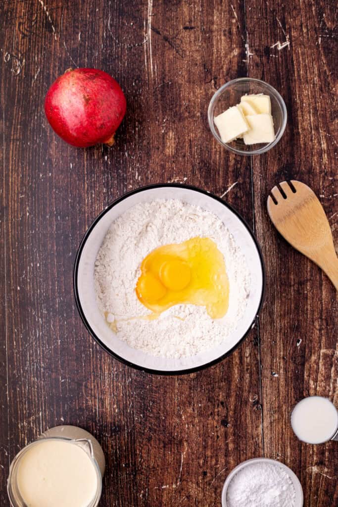 Mixing bowl filled with flour and cracked eggs in the center, surrounded by butter, milk, and other ingredients for making eggnog crepes.
