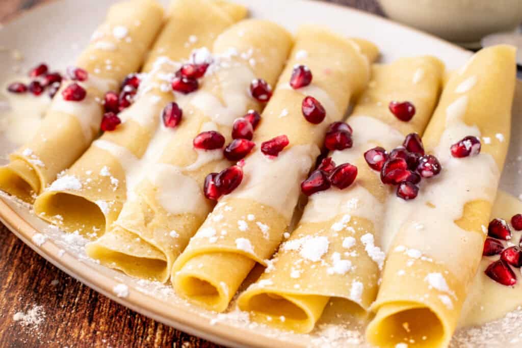 Angled view of golden eggnog crepes topped with creamy eggnog sauce and fresh pomegranate arils on a rustic serving plate.