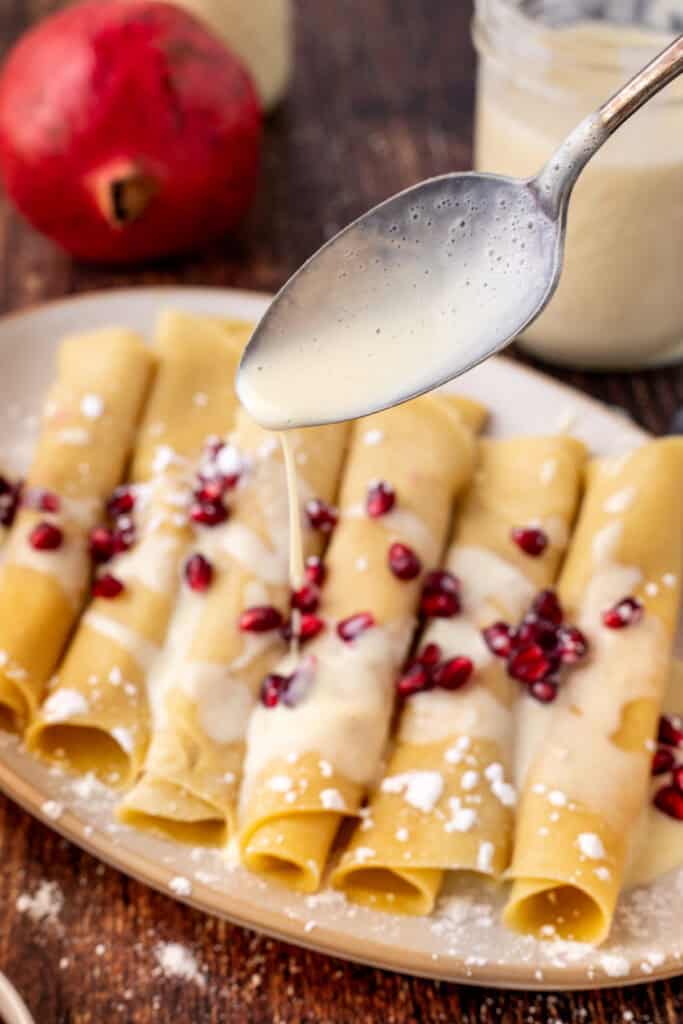 A close-up of a spoon drizzling creamy eggnog sauce over rolled crêpes topped with pomegranate seeds and a dusting of powdered sugar.