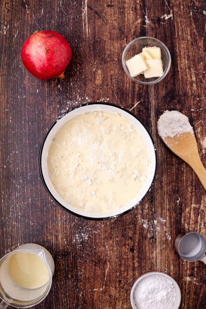 Mixing bowl with flour mixture and eggnog being combined to form crepe batter, surrounded by baking ingredients on a wooden table.