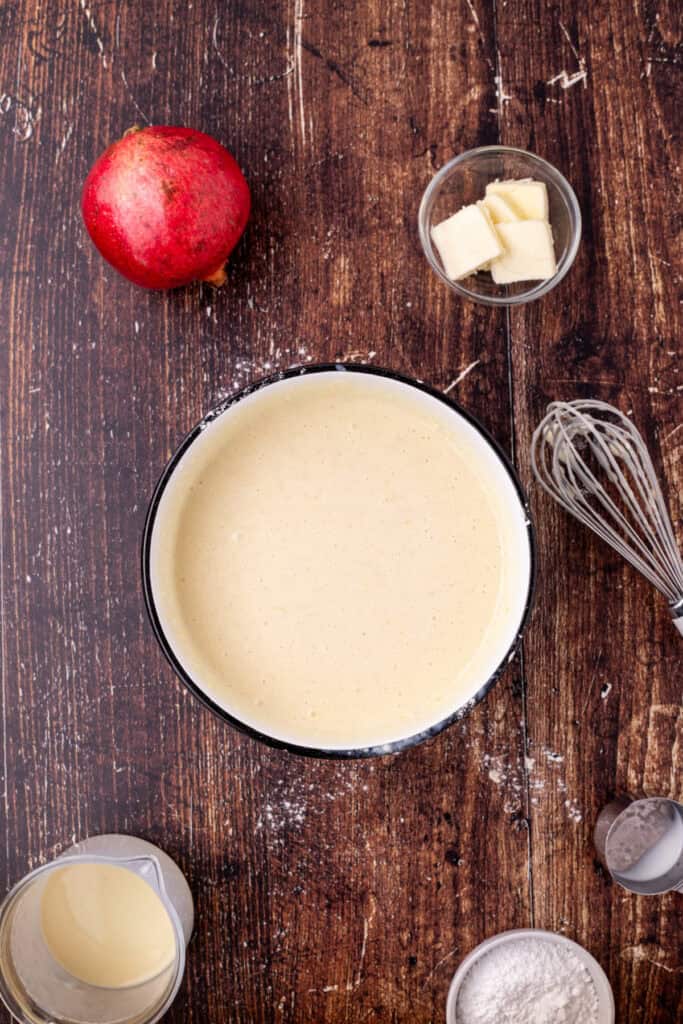 Smooth eggnog crepe batter in a bowl after whisking, with ingredients and utensils visible on a rustic wooden surface.