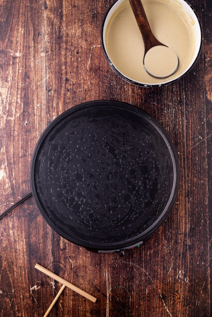 Crepe pan preheated and ready for cooking, with bowl of batter and wooden ladle nearby on a rustic wood table.