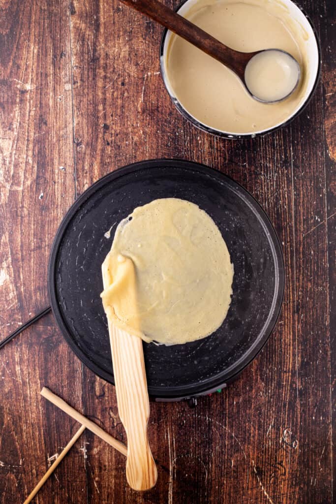 Partially cooked eggnog crepe being gently lifted with a wooden spatula from the pan.