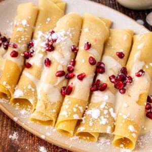 Square crop of eggnog crepes drizzled with eggnog sauce and topped with pomegranate arils, served on a beige plate over rustic wood.