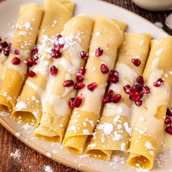 Square crop of eggnog crepes drizzled with eggnog sauce and topped with pomegranate arils, served on a beige plate over rustic wood.