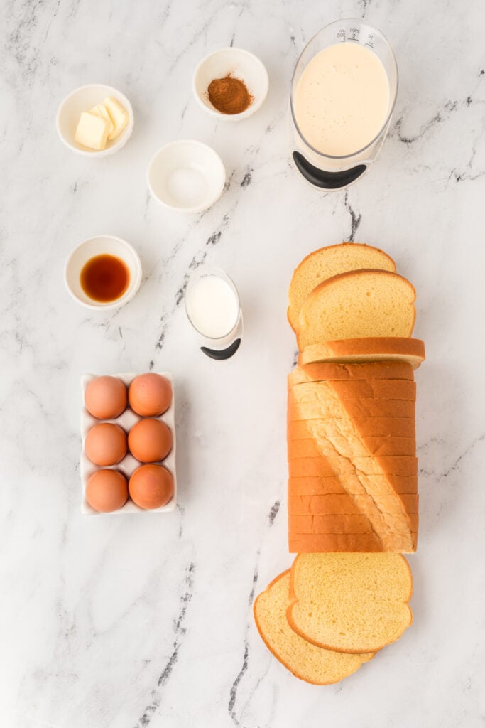Ingredients for Eggnog French Toast including eggs, milk, butter, vanilla, cinnamon, and sliced brioche bread on marble surface.