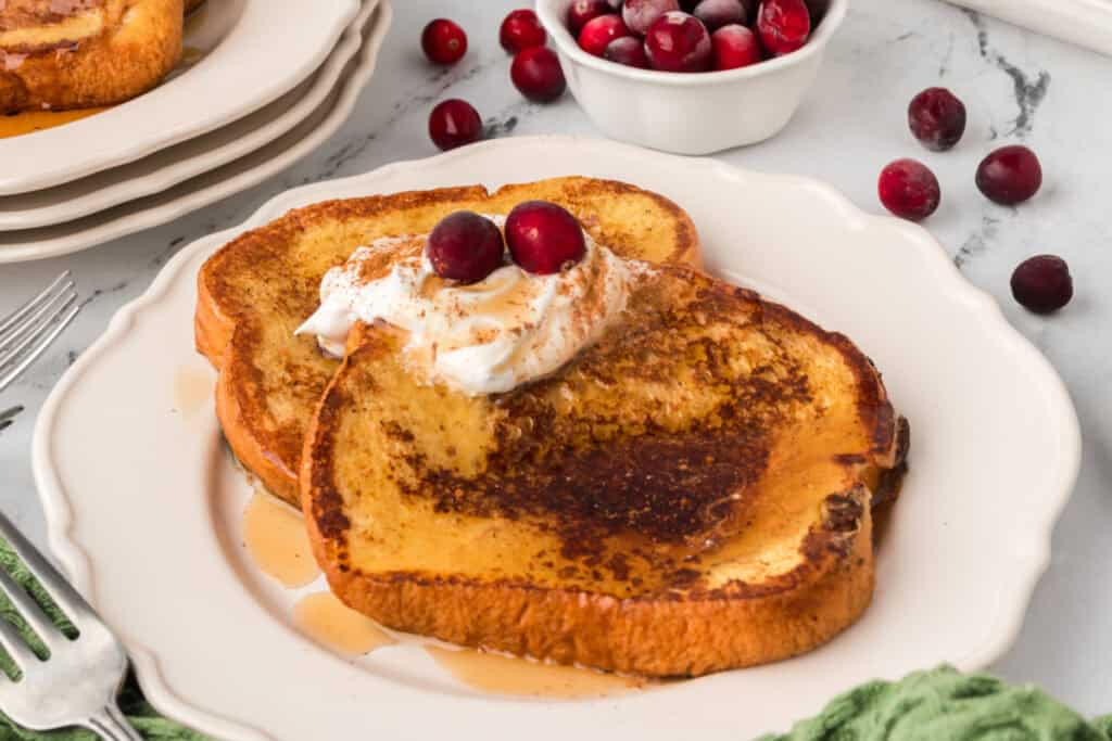 Two slices of Eggnog French Toast on a white plate topped with whipped cream, syrup, and cranberries, styled for the holidays.
