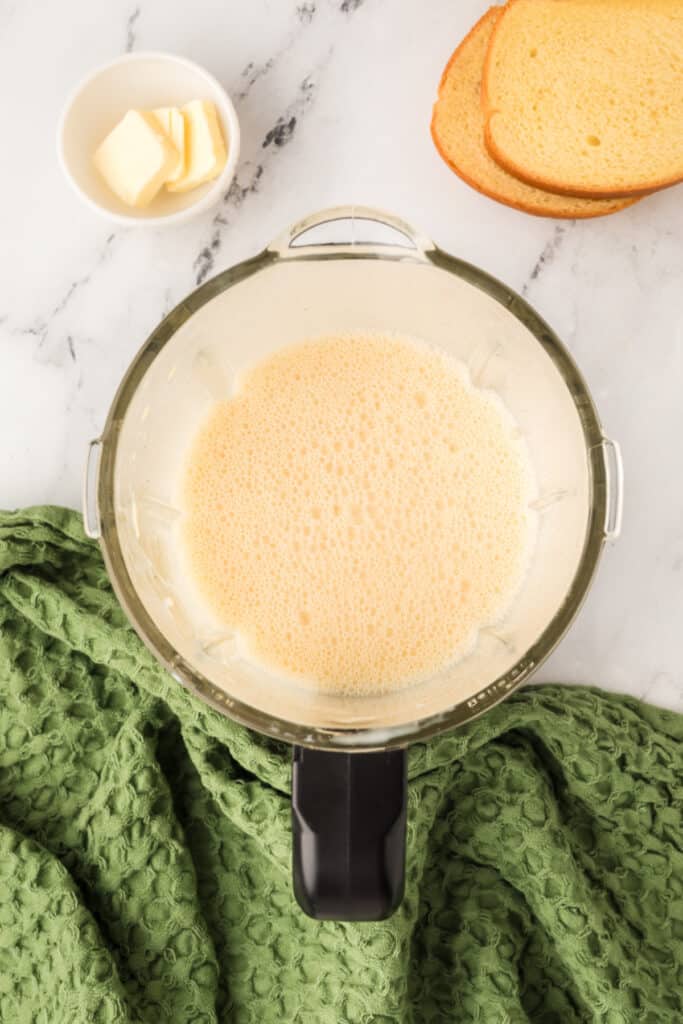 Blended eggnog mixture smooth and frothy in blender bowl beside green towel and bread slices.