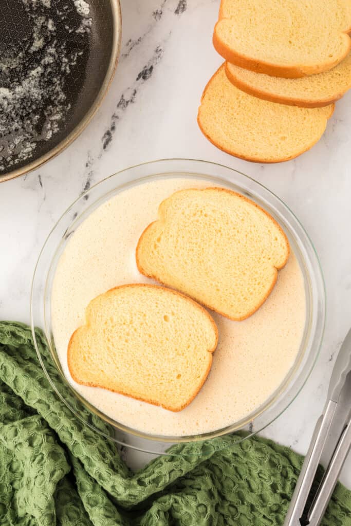 Slices of bread soaking in creamy eggnog mixture in a glass pie plate beside skillet.