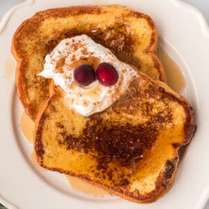 Square image of Eggnog French Toast topped with whipped cream, cinnamon, maple syrup, and cranberries on a white plate.
