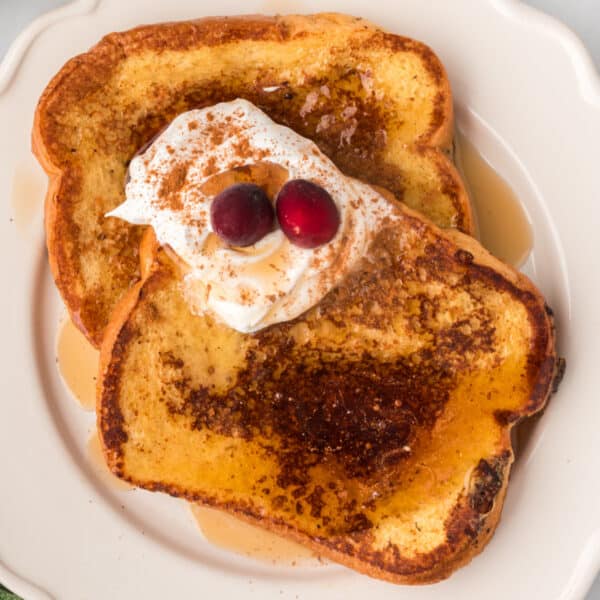 Square image of Eggnog French Toast topped with whipped cream, cinnamon, maple syrup, and cranberries on a white plate.