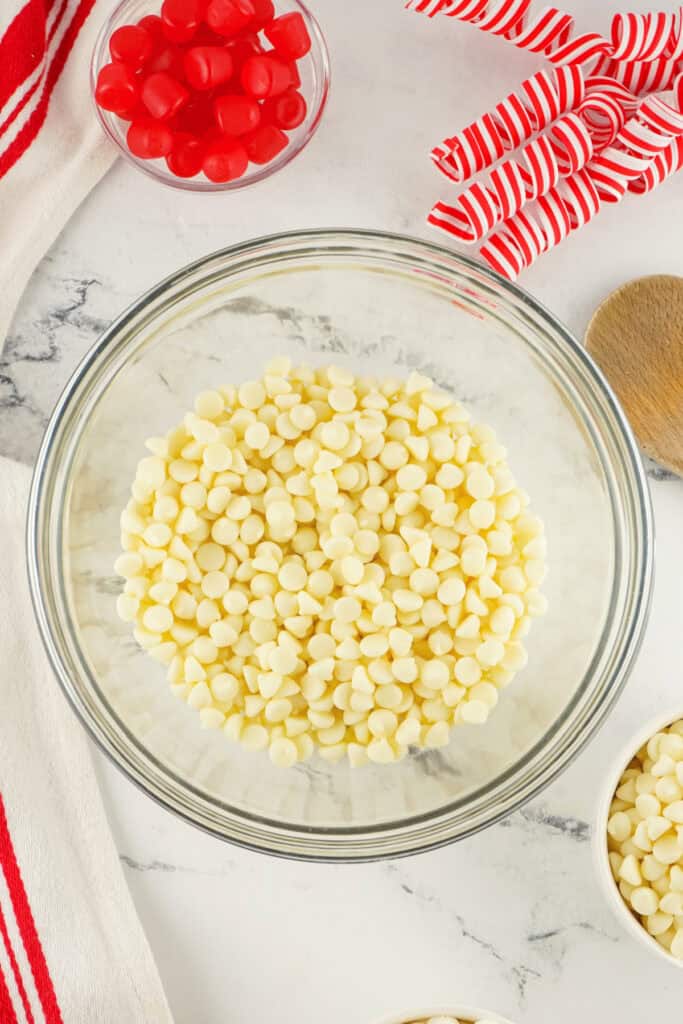 A glass bowl filled with white chocolate chips surrounded by red gumdrops and candy cane ribbon curls on a marble counter.