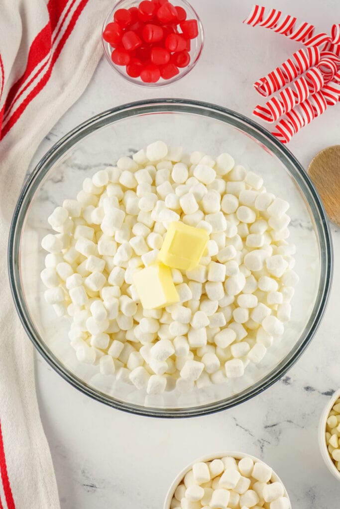 A large glass bowl of mini marshmallows with two pieces of butter on top, with red gumdrops and striped ribbon curls nearby.