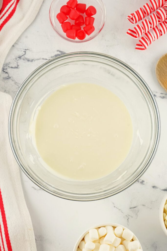 Melted white chocolate in a glass bowl on a marble surface with red gumdrops in a small dish beside it.