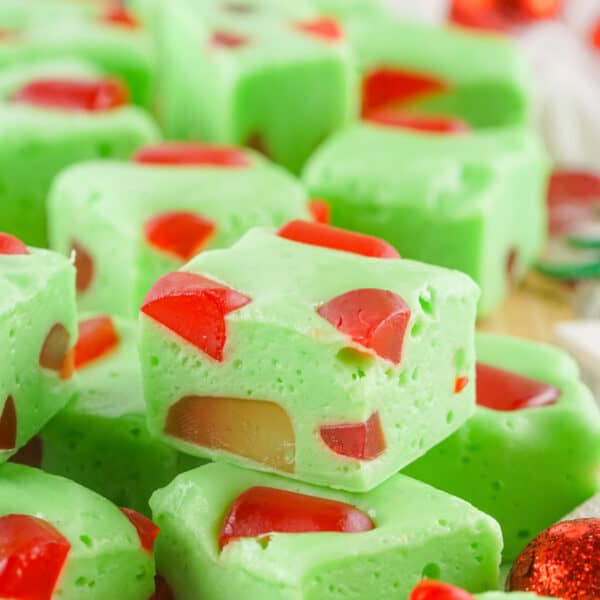 A square close up of green Grinch Christmas Nougat pieces filled with red gumdrops on a holiday themed surface.