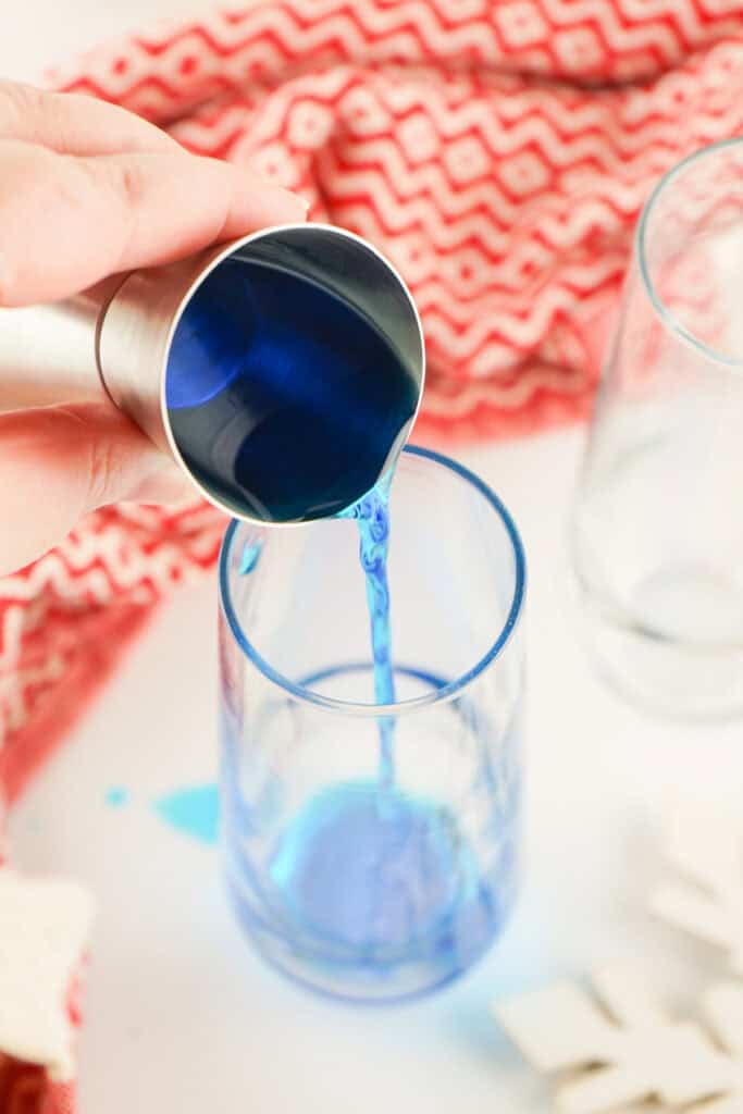 Blue curacao being poured from a jigger into a clear champagne glass with a red patterned cloth in the background