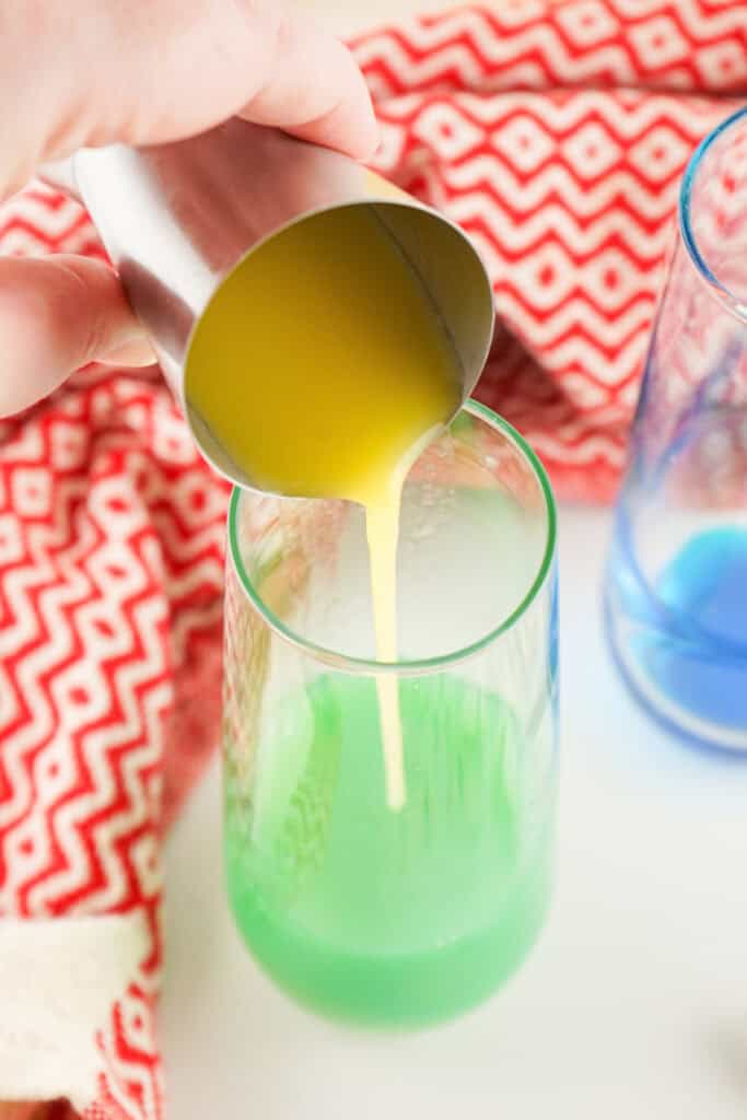 Orange juice being poured from a jigger into a glass of mixed blue curacao turning the drink bright green