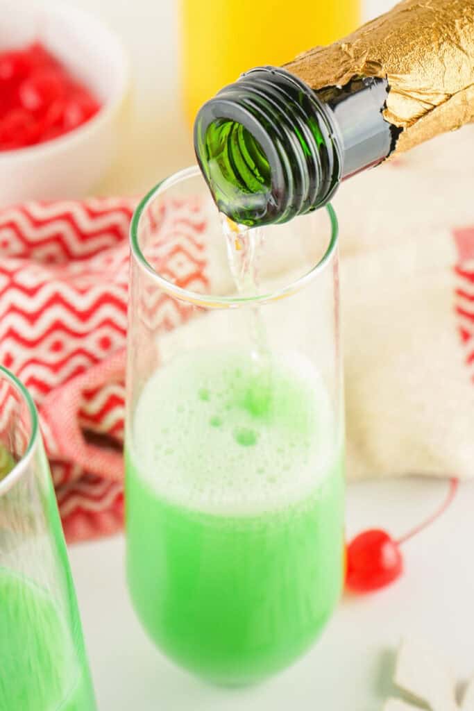 Champagne being poured from a green bottle into a glass of green mimosa mixture with cherries nearby