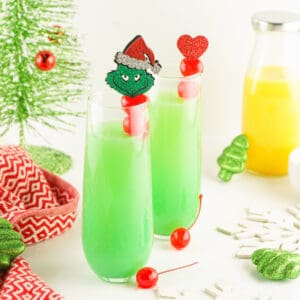 Two bright green Grinch Mimosas with red cherry toppers surrounded by holiday decor and a bottle of orange juice