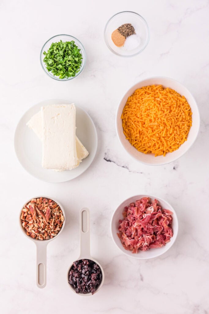 Overhead view of ingredients for Holiday Cheese Ball, including cream cheese, shredded cheddar, prosciutto, pecans, dried cherries, parsley, and seasonings on a marble surface.