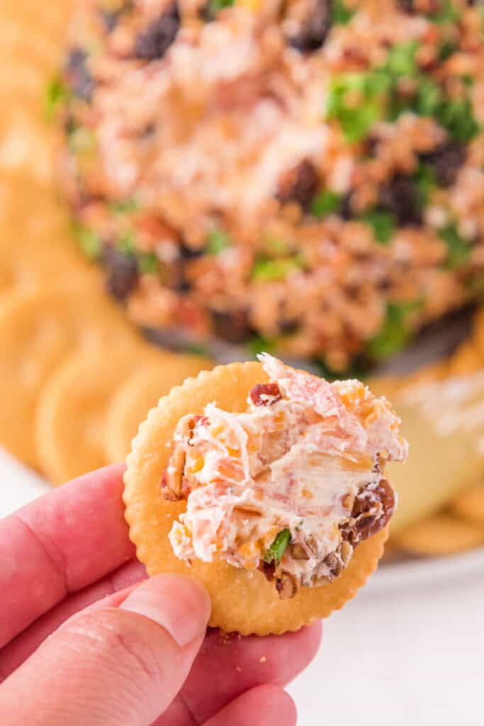 Hand holding a cracker topped with a bite of creamy Holiday Cheese Ball.