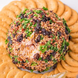 Square crop of Holiday Cheese Ball coated in pecans, cherries, and parsley, surrounded by crackers on a serving plate.