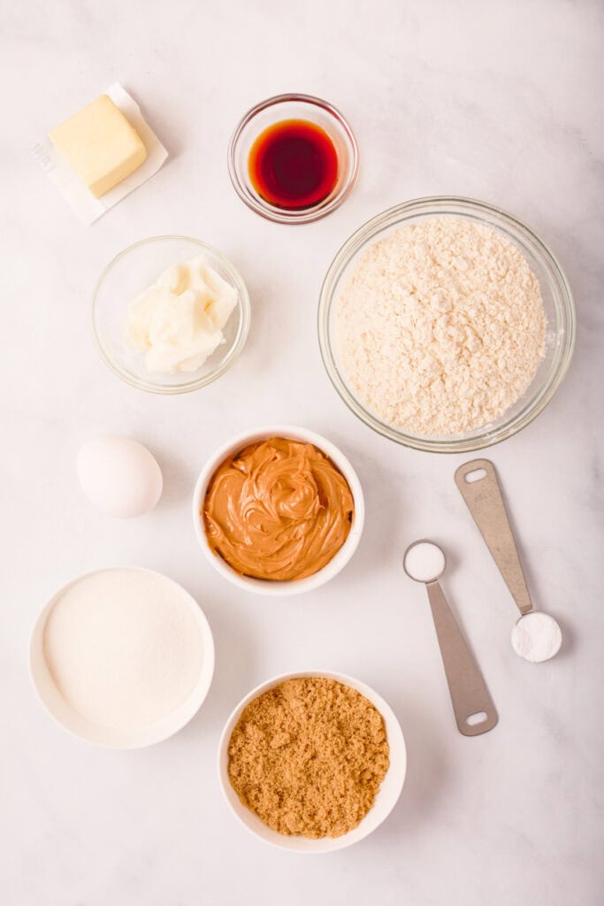 Overhead view of measured peanut butter cookie ingredients including butter, shortening, peanut butter, sugars, flour, egg, vanilla, salt, and baking soda arranged on a white surface.