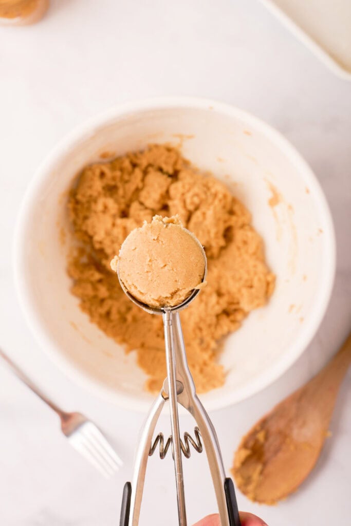 Cookie scoop holding a portion of peanut butter dough above the mixing bowl, showing the dough’s soft texture before baking.