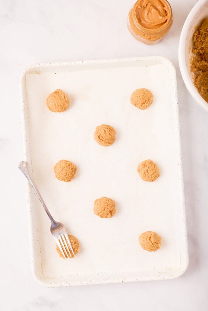 Baking sheet lined with parchment paper showing scoops of peanut butter cookie dough spaced apart with a fork ready for crisscrossing.