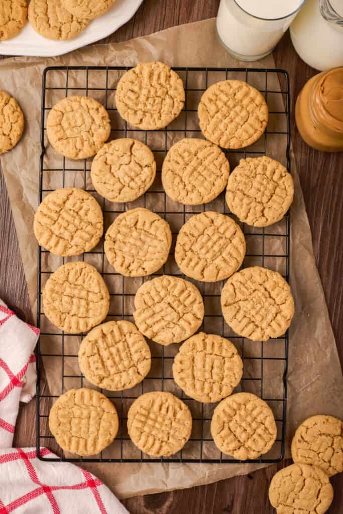 Peanut butter cookies cooling on a wire rack with parchment underneath, surrounded by milk and a jar of peanut butter.