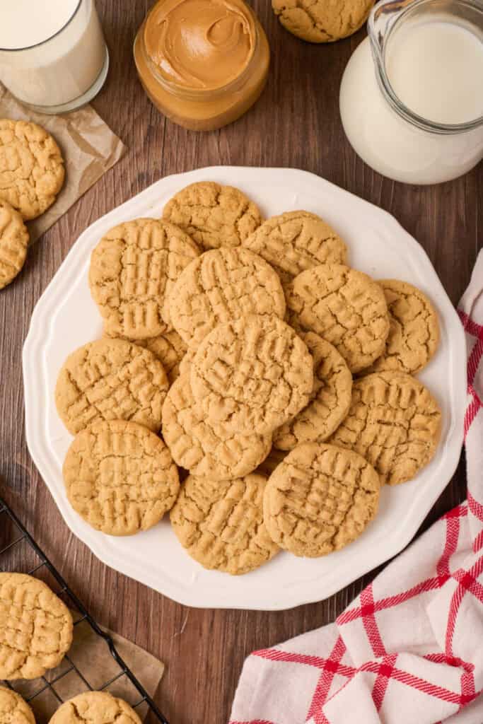 Plate piled high with peanut butter cookies beside a jar of peanut butter and glasses of milk on a wooden table.
