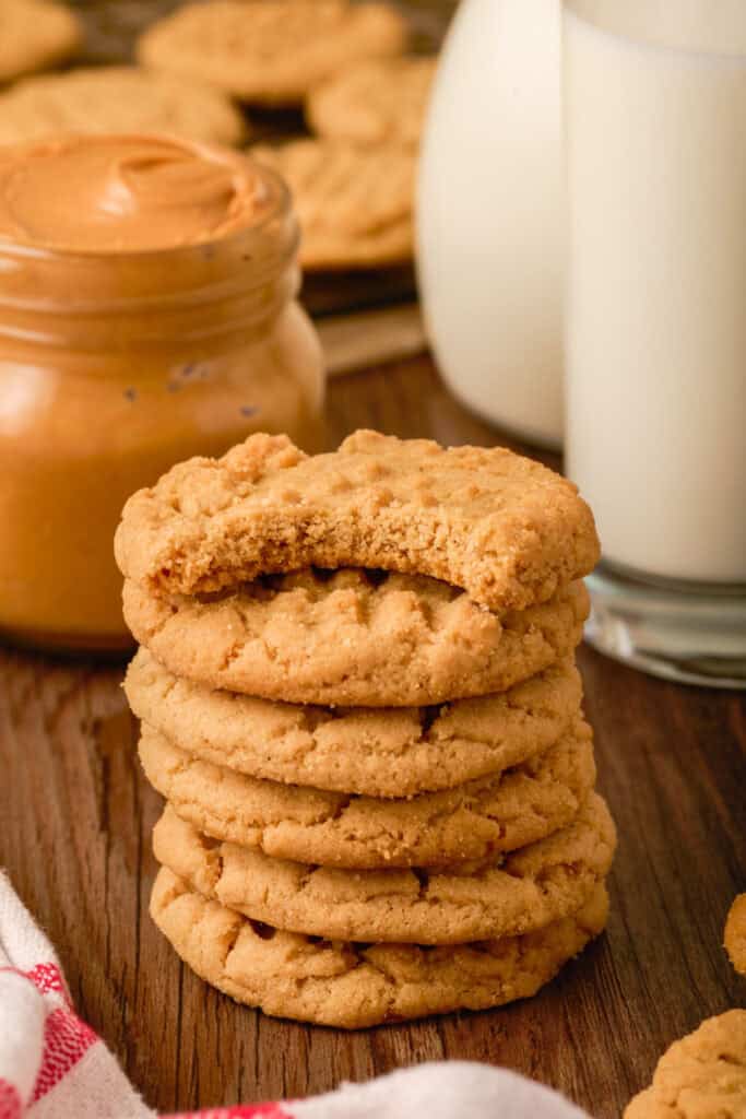 Stack of peanut butter cookies with a bite taken from the top cookie, sitting beside milk and peanut butter.
