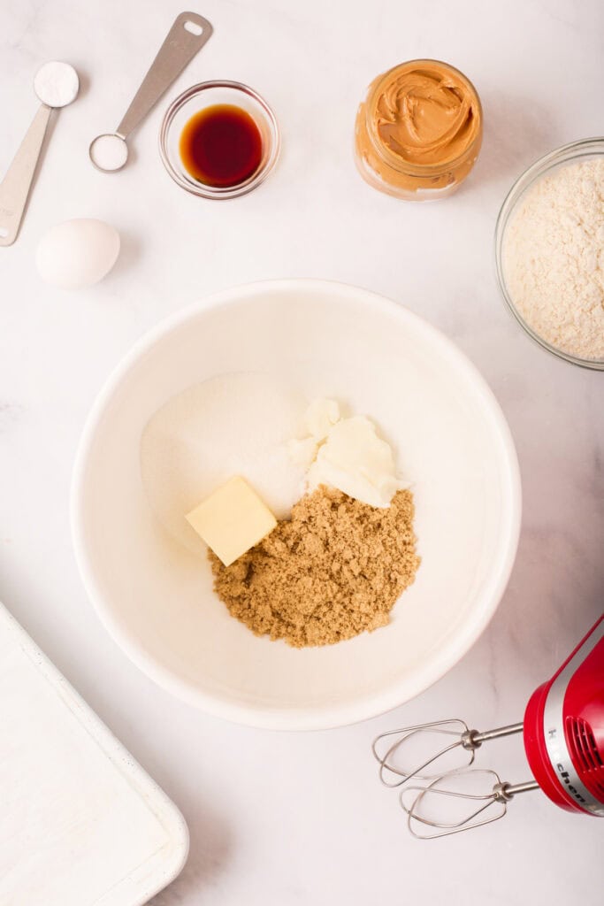 Mixing bowl with butter, shortening, brown sugar, and granulated sugar before creaming, surrounded by additional cookie ingredients and a red hand mixer.