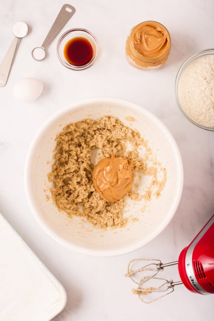 Peanut butter added to the creamed butter and sugar mixture in a white bowl with remaining ingredients and tools visible on the counter.