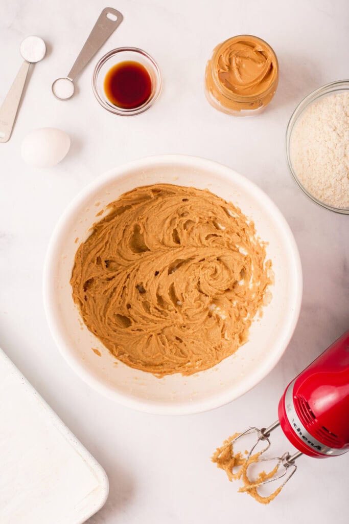 Peanut butter cookie dough after being mixed smooth and creamy in a white bowl using a red hand mixer.