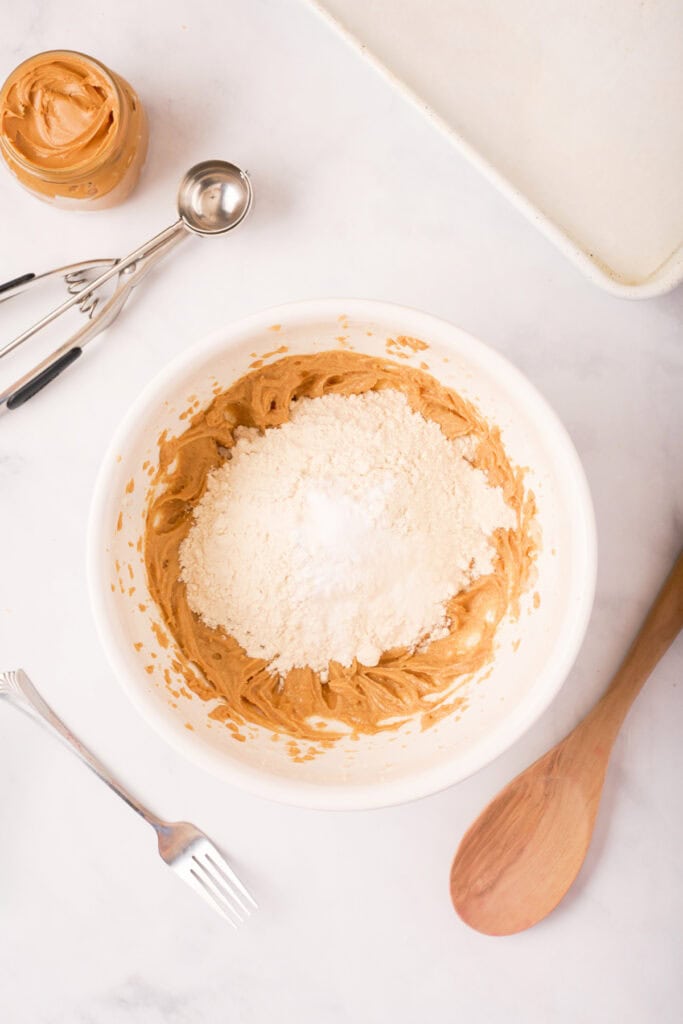 Flour, baking soda, and salt added on top of the peanut butter mixture in a bowl, ready to be stirred in with a wooden spoon.