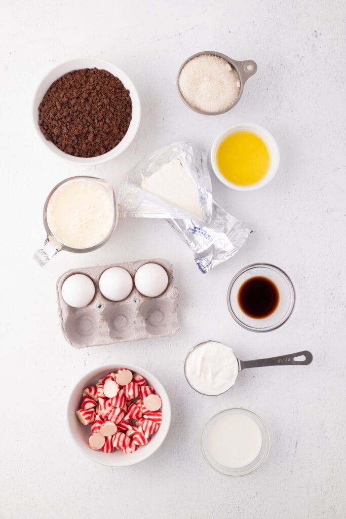 Overhead view of ingredients for peppermint eggnog cheesecake, including cream cheese, eggs, graham crumbs, melted butter, sugar, vanilla, eggnog, sour cream, milk, and peppermint candy cane Kisses arranged on a white surface.