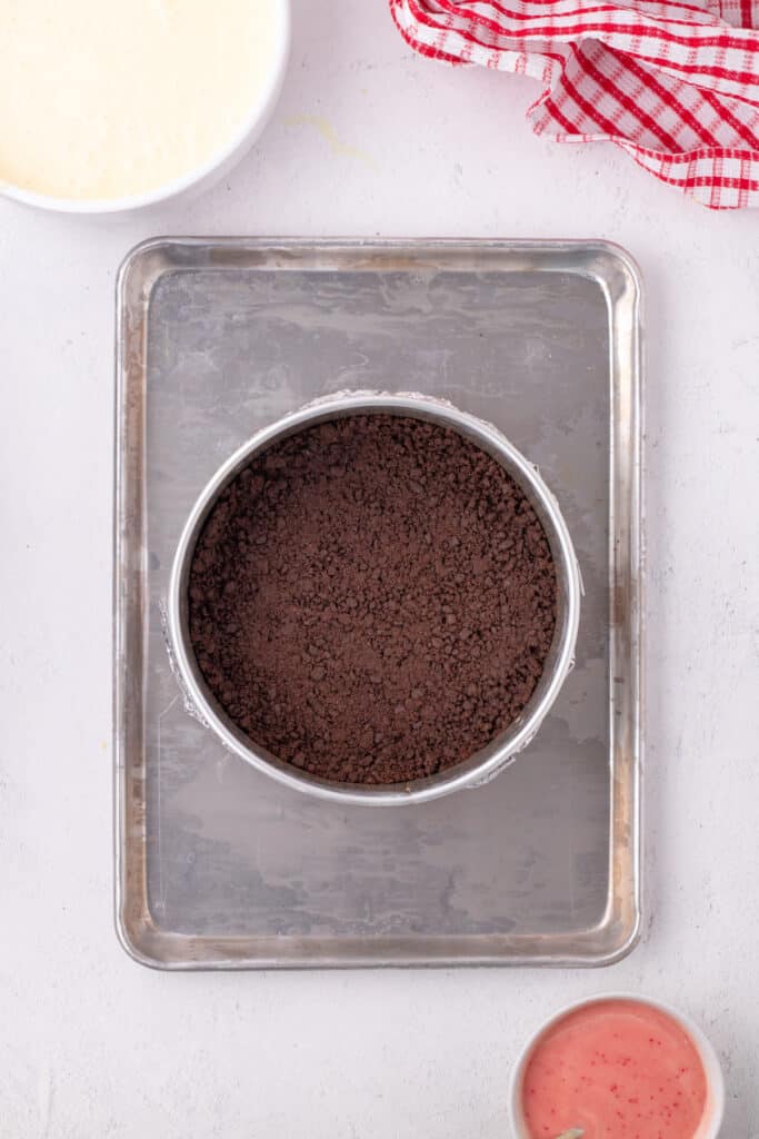 Springform pan with baked chocolate crust set on a sheet pan, with bowls of plain cheesecake batter and pink peppermint batter nearby.