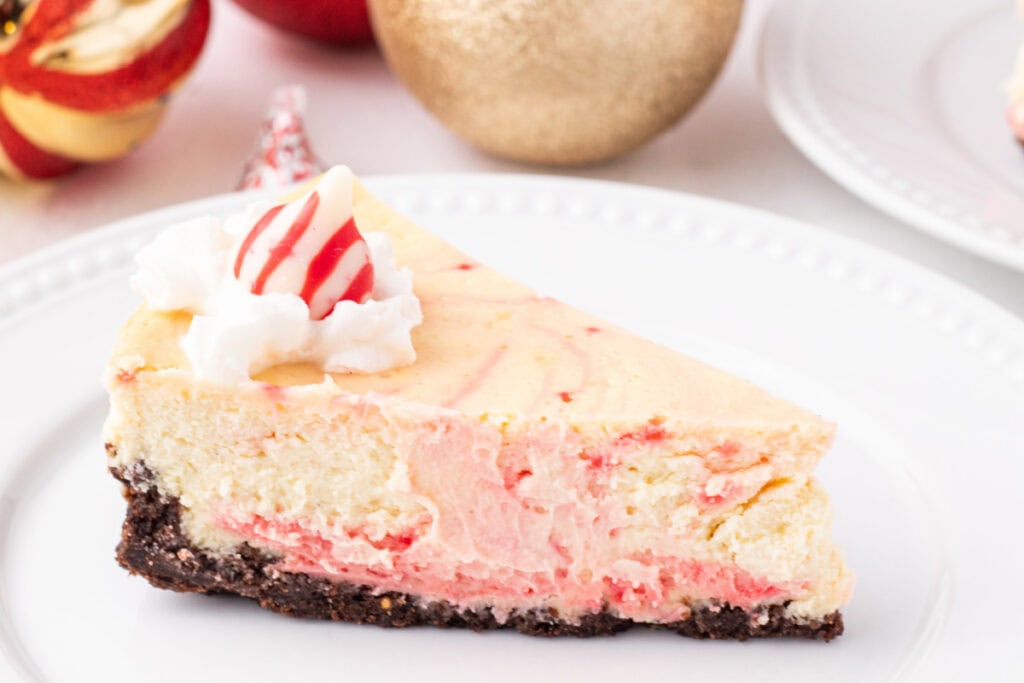 Slice of peppermint eggnog cheesecake on a white plate, showing the pink peppermint layer, creamy filling, chocolate crust, and a whipped cream and peppermint Kiss garnish.
