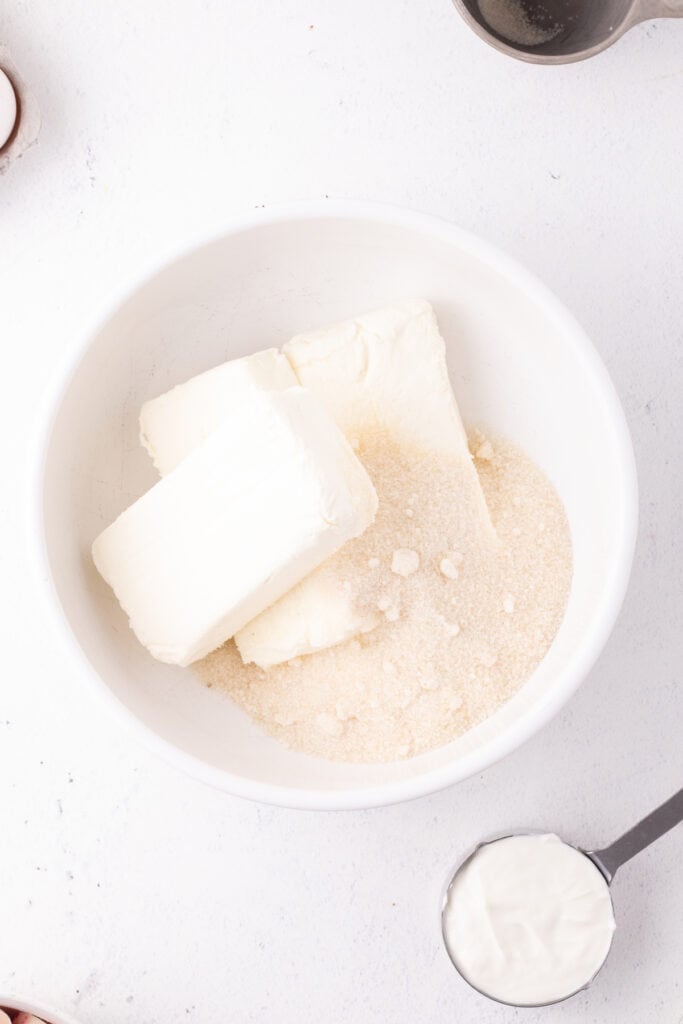 Cream cheese blocks and granulated sugar placed together in a white mixing bowl before blending for the cheesecake filling.