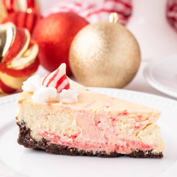 Close view of a slice of peppermint eggnog cheesecake on a white plate with holiday ornaments in the background, topped with whipped cream and a peppermint Kiss.