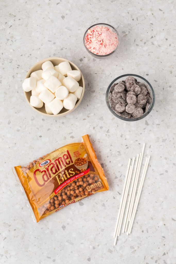 Ingredients laid out on a countertop, including a bowl of marshmallows, a bowl of crushed peppermints, a bowl of dark chocolate wafers, a bag of caramel bits, and several lollipop sticks.