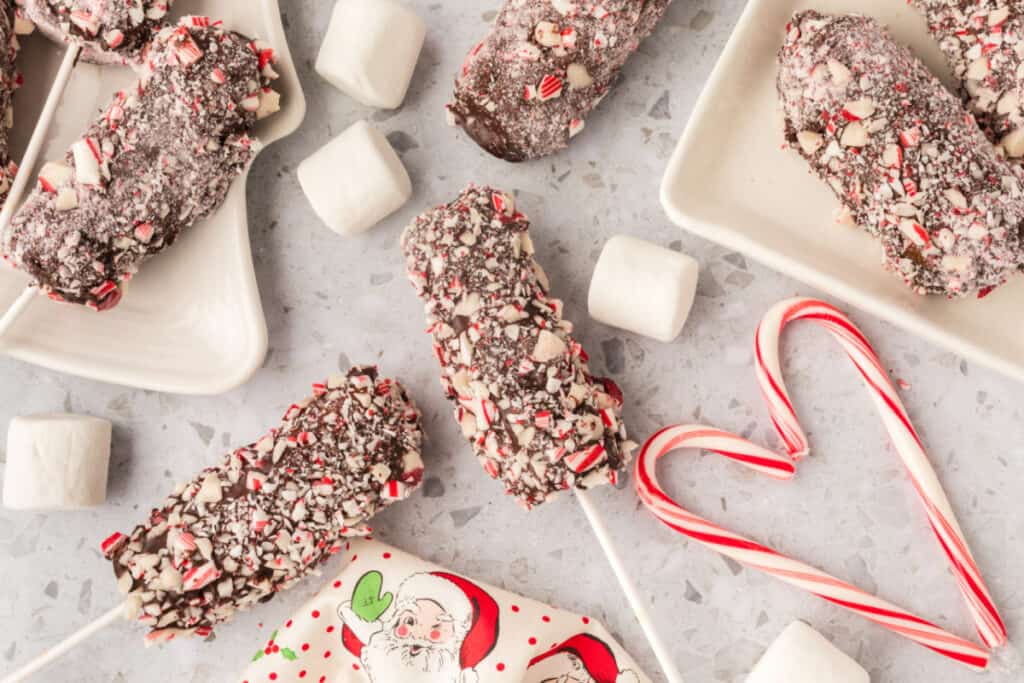 Multiple peppermint coated marshmallow wands scattered on plates and the countertop with loose marshmallows and candy canes nearby.