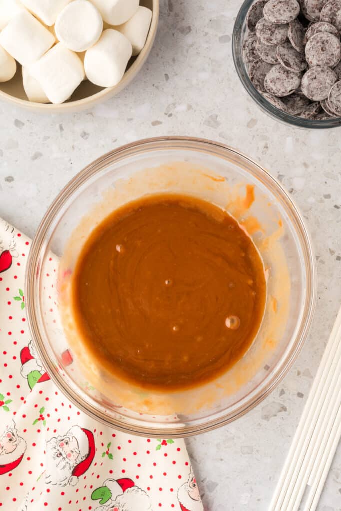 A glass bowl filled with melted caramel, surrounded by marshmallows, chocolate wafers, and lollipop sticks on a light countertop.