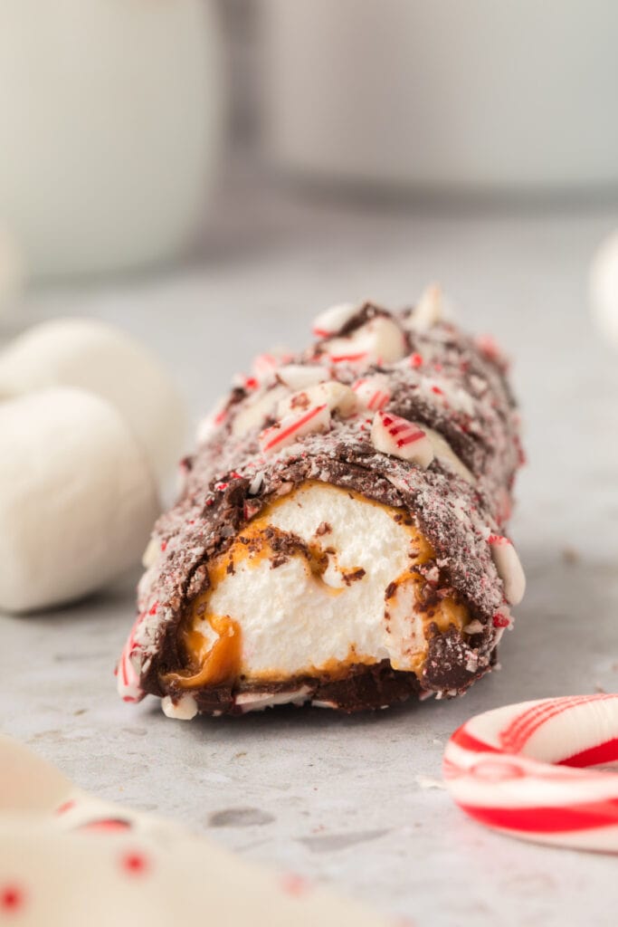 A single peppermint coated marshmallow wand with a bite removed resting on the countertop with loose marshmallows and candy canes nearby.