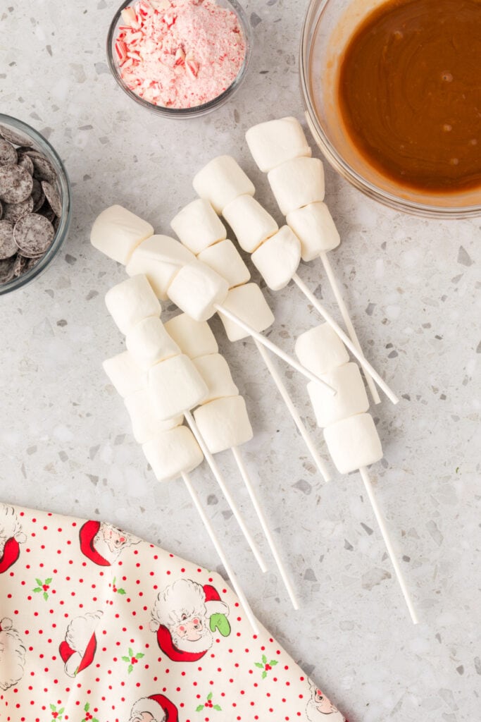 Several lollipop sticks each holding three marshmallows, arranged on a countertop beside melted caramel and crushed peppermint.