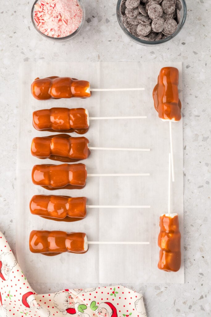 Marshmallow wands coated in caramel resting on parchment paper, with bowls of chocolate wafers and crushed peppermint nearby.