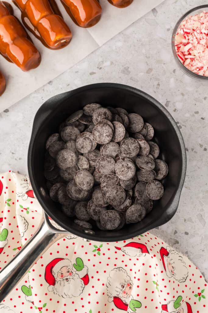 A saucepan filled with dark chocolate melting wafers sitting next to caramel coated marshmallow wands.