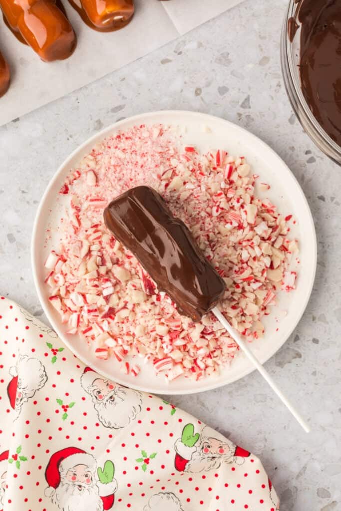 A caramel coated marshmallow wand dipped in melted chocolate resting on a plate of crushed peppermint.